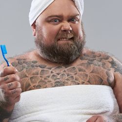 Close up of bearded tattooed man standing against the grey background and biting his lower lip while holding a toothbrush in right hand