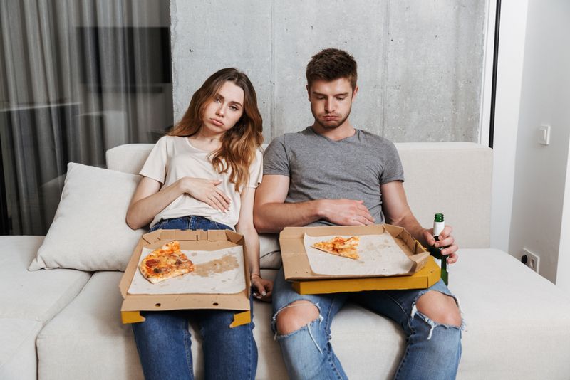 Man and woman sitting on white sofa with pizza leftovers, looking very full.
