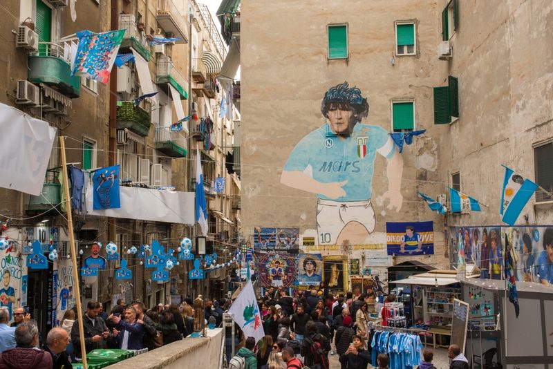 A busy city streety in Naples, Italy, with a graffiti portrait of Maradona on the wall.