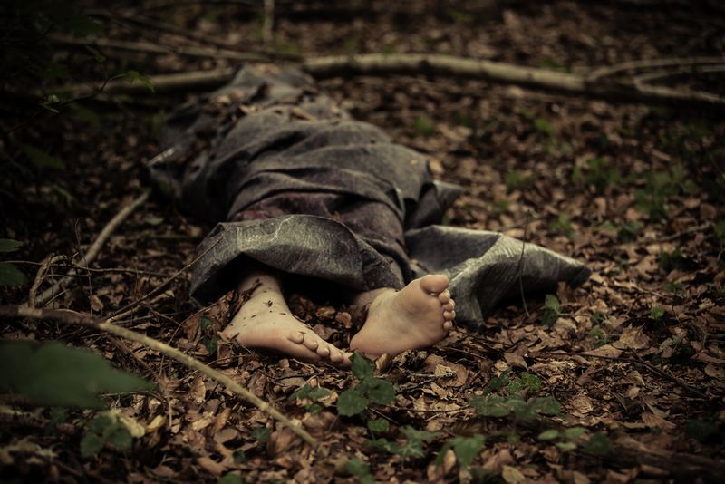 Person wrapped in grey blanket on forest floor, feet protruding from bottom of blanket