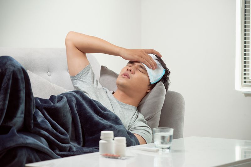 man lying ill on sofa with cold compress on forehead and table with water, medicines and thermometer