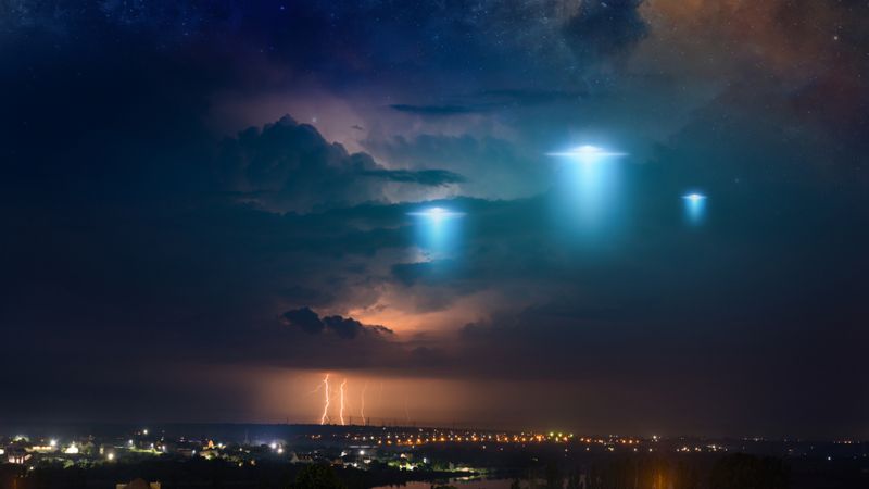 Extraterrestrial aliens spaceship fly above small town, ufo with blue spotlights in dark stormy sky. Elements of this image furnished by NASA